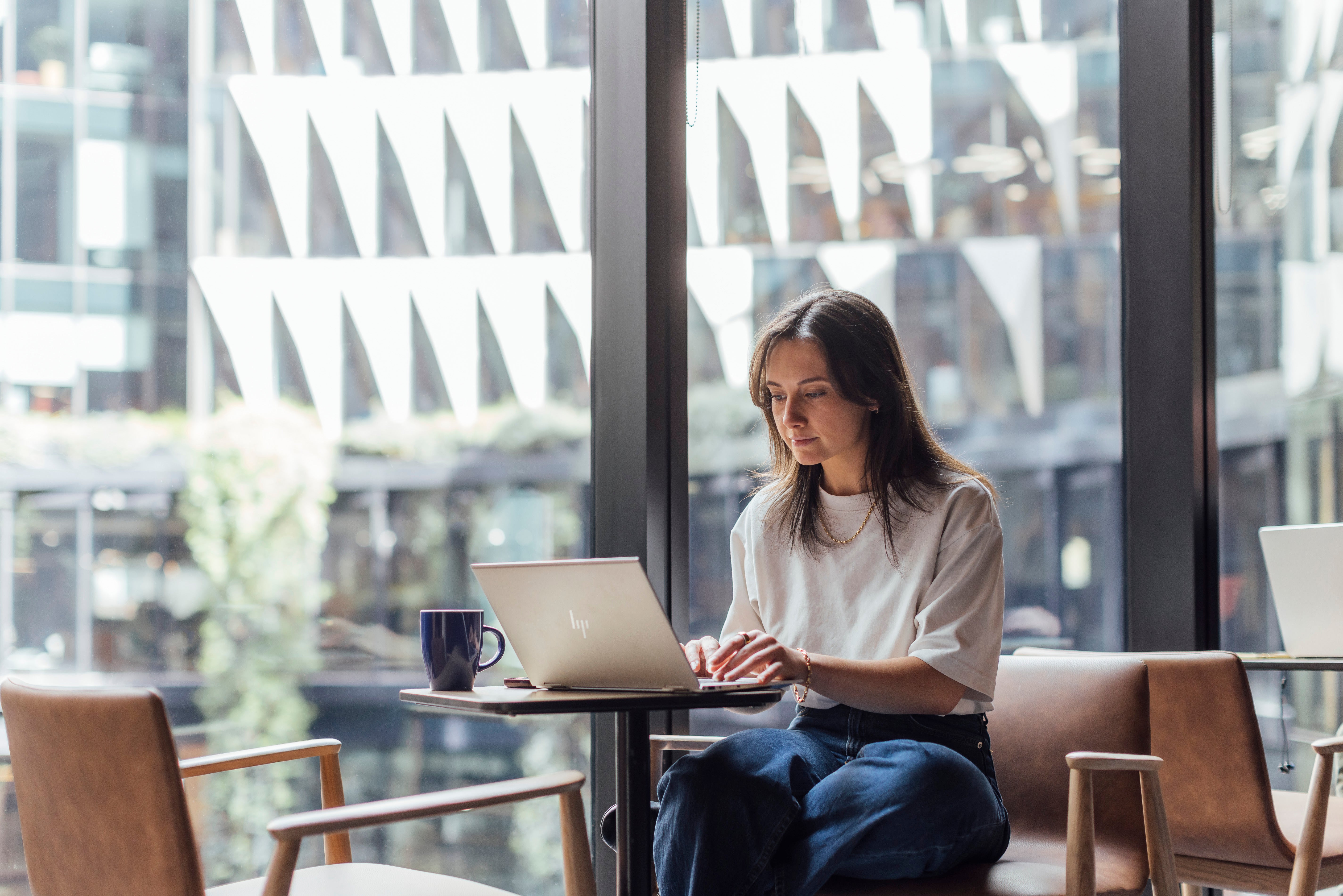 A person sits at a table by a large window, typing on a laptop. A blue mug sits on the table next to the laptop. The background shows a modern building with geometric patterns.