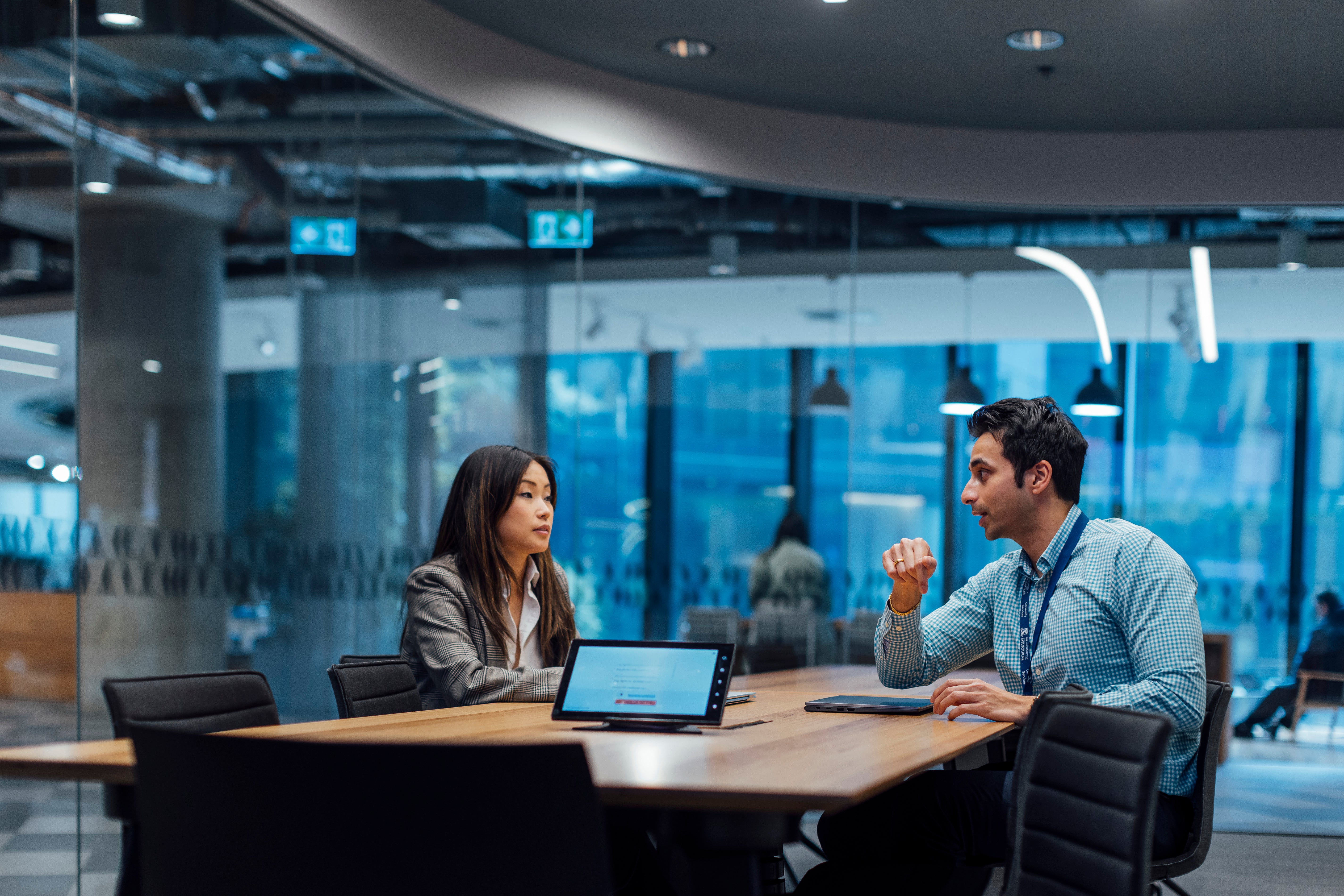 Two people, one facing the camera and the other in profile, sit at a conference table discussing something. A tablet is on the table between them.