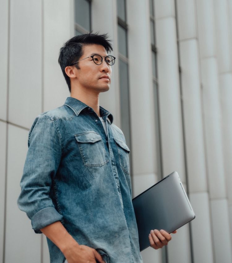 Man with a laptop standing outside a building, gazing thoughtfully into the distance.