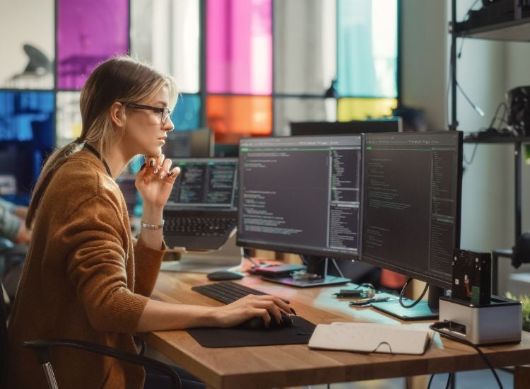 A woman working at a desk with two computer monitors, focused on her tasks.