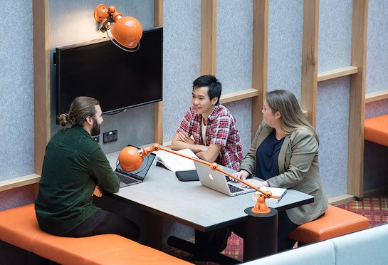An aerial shot of 3 people having a conversation at a communal table, with laptops and textbooks open in front of them.