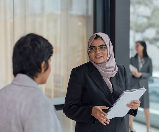A woman wearing glasses and a headscarf speaks to her colleague, in the foreground, whilst on campus.