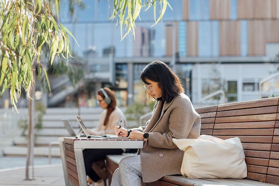 A dark-haired woman sits at a public bench. She is making notes while focused on a laptop open on the table in front of her.