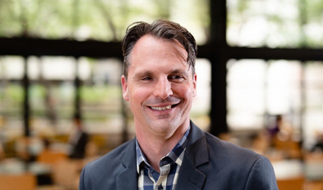 Professor Toby Murray is smiling while looking slightly to the right, with a blurred university building interior behind him.