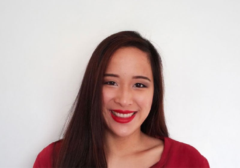 A portrait style photo of Miranda, a woman with dark hair and a bright red lip, looking to camera and smiling. 