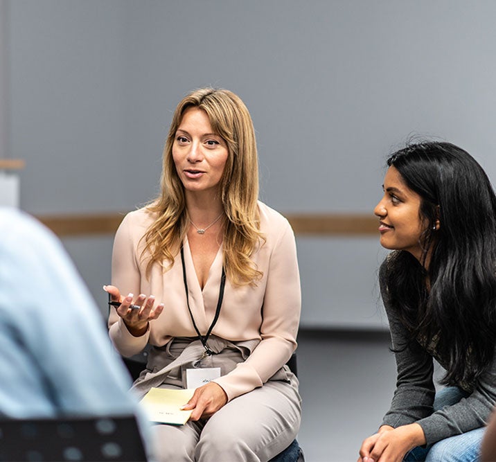A woman speaks while holding papers in a group discussion. Two other people sit and listen attentively.
