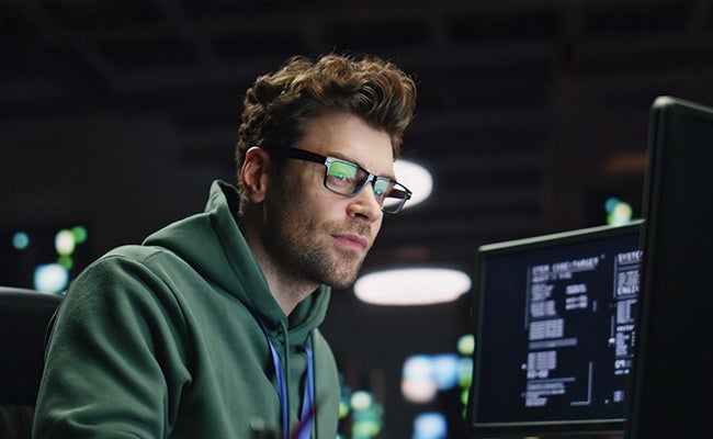 A man with glasses on sits at a desk with a dark background and multiple screens. He's typing while looking at one screen.