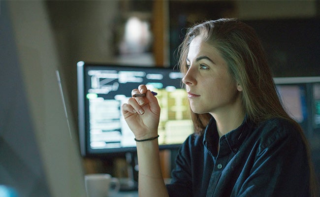 A woman in sits side on with a computer screen behind her, looking at the screen in front of her while holding a pen.
