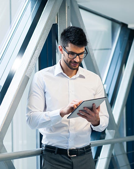 A man in a white shirt stands in an office corridor with a window behind. He's tapping the ipad he's holding in front of him.