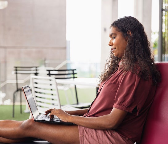 A woman with dark hair reclines on a plum couch with a laptop in her lap. She appears focused on what she's typing.