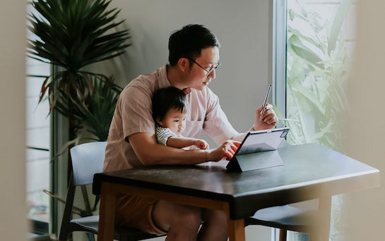 A man sits at a table with a child in his lap. He is tapping on a tablet while looking at his stylus.