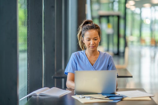A woman in pale blue medical scrubs sits in an office foyer and works on her laptop. A textbook is open on the table next to her.