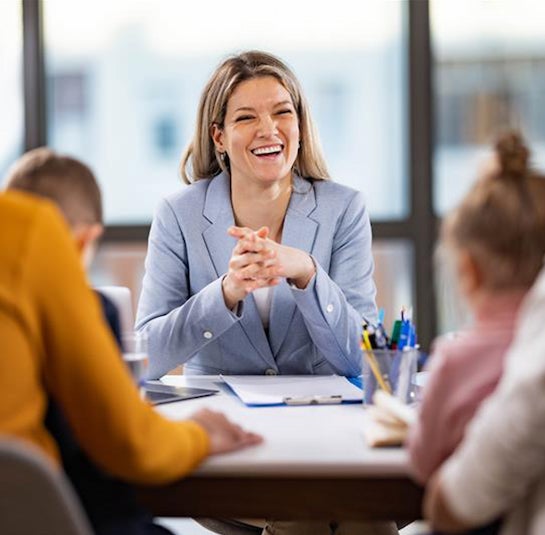 A blonde woman in a blue blazer is seated, smiling as she addresses adults and children partially shown in the foreground.