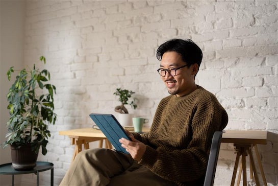 A man with dark hair and glasses is looking a digital tablet and smiling. Behind him are pot plants and a bright textured wall.