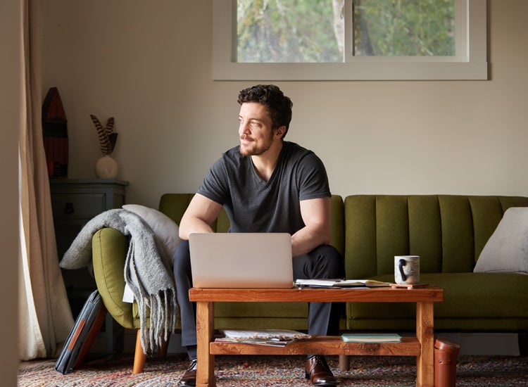 A bearded man on a green sofa looking out a window. A laptop and a small speaker on the coffee table.