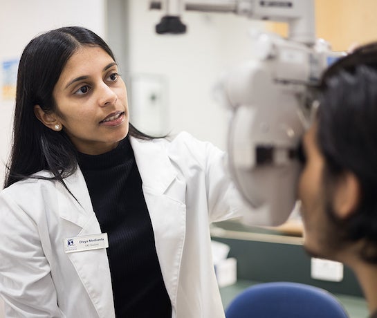 A dark-haired woman in a white lab coat uses an eye testing machine on a patient. She is wearing a University of Melbourne name badge.