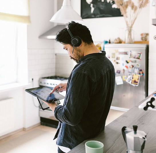 A man with dark hair and headphones is in a kitchen, interacting with a digital tablet while on a virtual call.