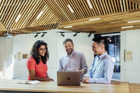 3 people are in a modern university facility, looking at a laptop on the table in front of them and smiling.